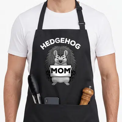Hedge animal apron - Cartoon hedgehog holding a white sign with black text reading MOM under arching HEDGEHOG letters — shown on a black cotton canvas apron against a white background
