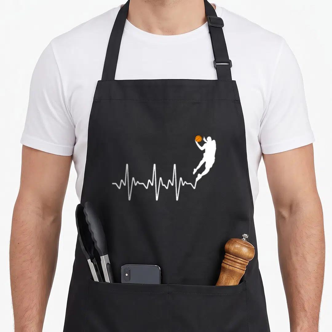 Funny player basketball coach apron - White heartbeat line morphing into jumping basketball player silhouette holding — shown on a black cotton canvas apron against a white background