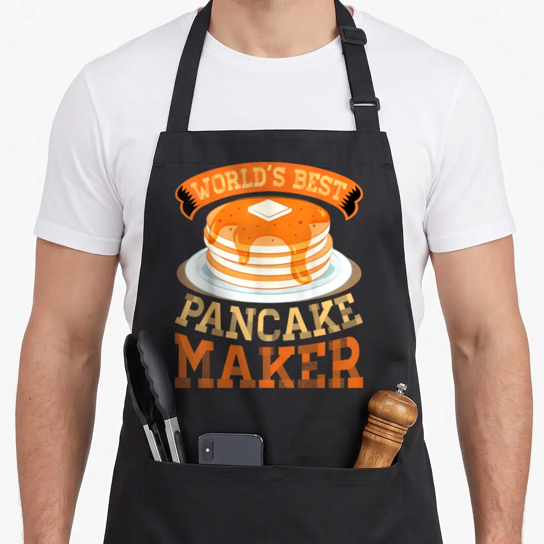 Funny maker pancake apron - Cartoon stack of pancakes under orange ribbon reading WORLD'S BEST PANCAKE MAKER — shown on a black cotton canvas apron against a white background