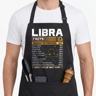 Libra fact birthday apron - White and yellow nutrition label style text listing Libra traits like savage and loyal — shown on a black cotton canvas apron against a white background