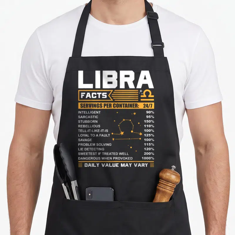 Libra fact birthday apron - White and yellow nutrition label style text listing Libra traits like savage and loyal — shown on a black cotton canvas apron against a white background
