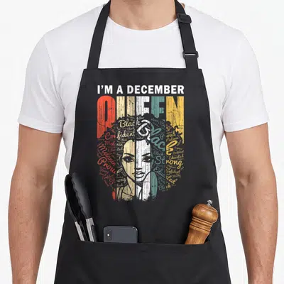 Queen december birthday apron - I'M A DECEMBER QUEEN text with colorful collage face and empowering words like strong — shown on a black cotton canvas apron against a white background