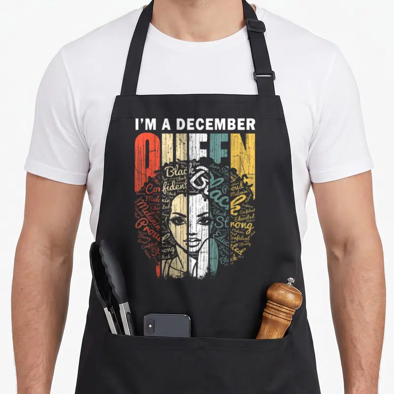 Queen december birthday apron - I'M A DECEMBER QUEEN text with colorful collage face and empowering words like strong — shown on a black cotton canvas apron against a white background