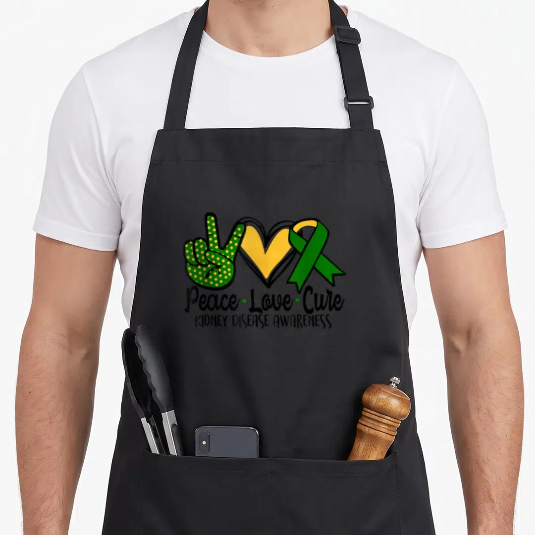 March ribbon apron - Green peace sign hand, yellow heart, green ribbon, and text Peace Love Cure Kidney Disease — shown on a black cotton canvas apron against a white background