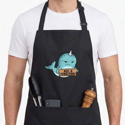 Whale unicorn apron - Grumpy blue cartoon narwhal holding brown sign with text NOT A UNICORN — shown on a black cotton canvas apron against a white background