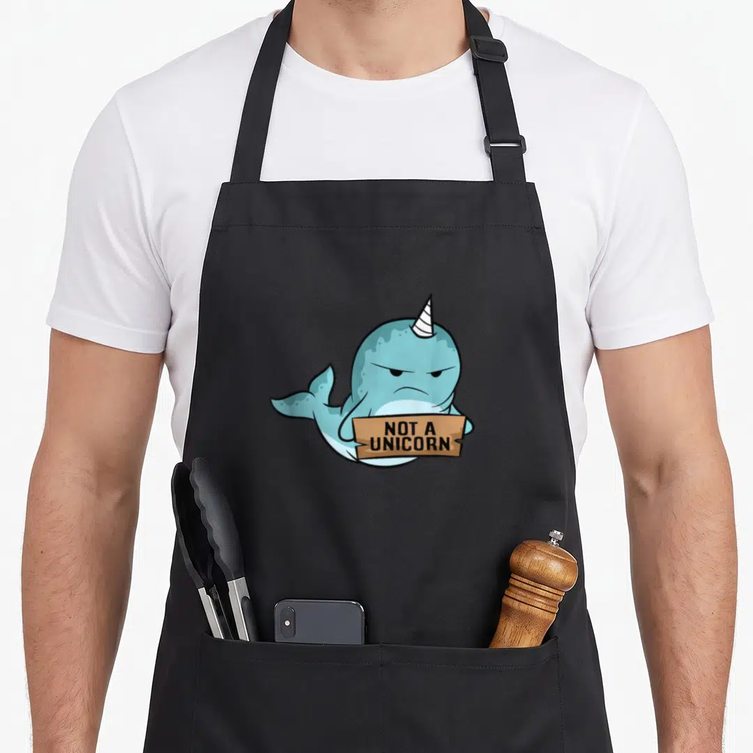 Whale unicorn apron - Grumpy blue cartoon narwhal holding brown sign with text NOT A UNICORN — shown on a black cotton canvas apron against a white background