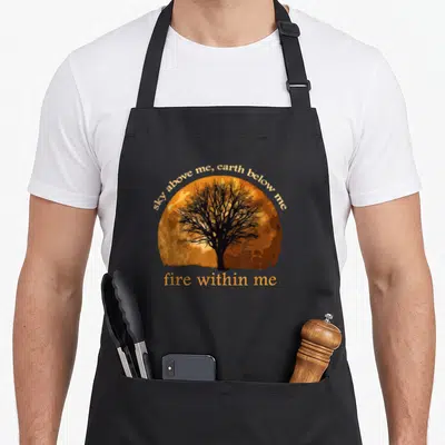 Fire pagan apron - Black tree silhouette on orange sun with sky above me earth below me fire within me text — shown on a black cotton canvas apron against a white background
