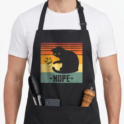 King black cat apron - Grey cat silhouette knocking over crown with nope text on retro stripes — shown on a black cotton canvas apron against a white background