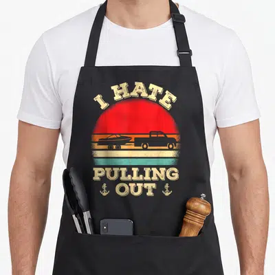 Hate boat apron - Retro truck boat silhouette sunset design with I HATE PULLING OUT text and two anchors — shown on a black cotton canvas apron against a white background