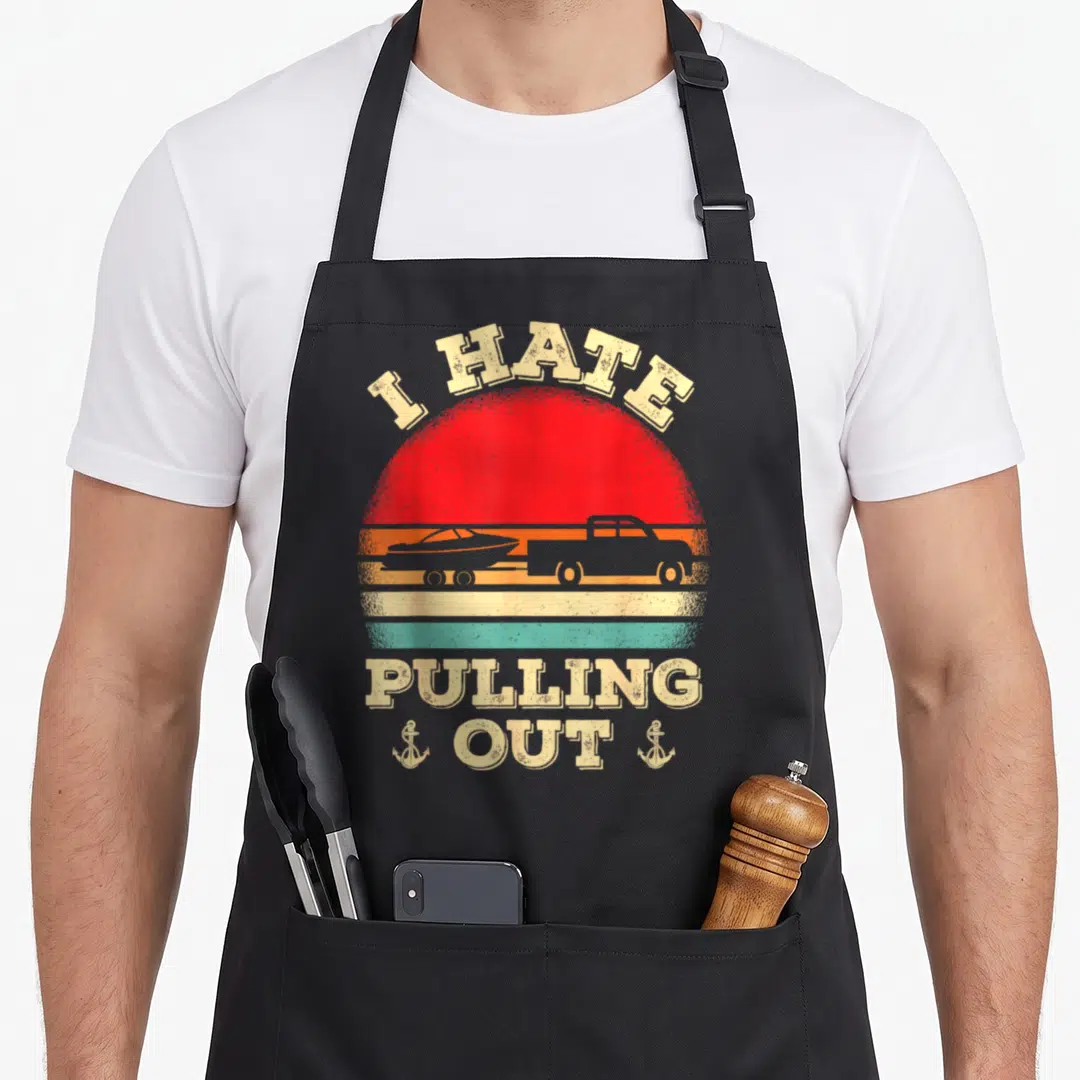 Hate boat apron - Retro truck boat silhouette sunset design with I HATE PULLING OUT text and two anchors — shown on a black cotton canvas apron against a white background