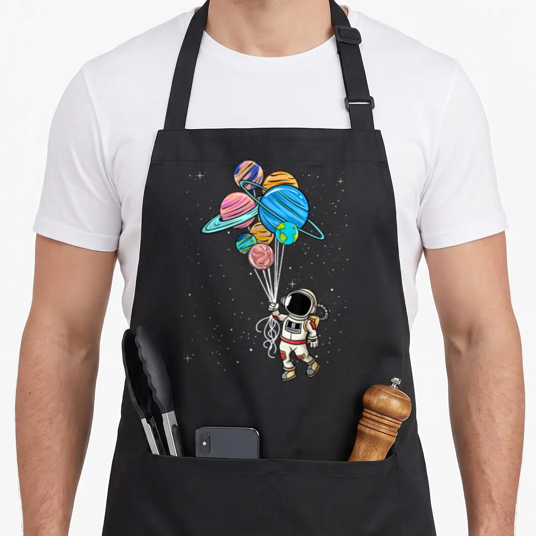 Planet balloon apron - Cartoon astronaut holding seven striped planetary balloons in space with scattered white stars — shown on a black cotton canvas apron against a white background
