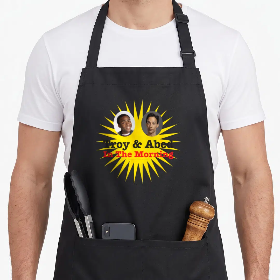 Troy community squad apron - Two circular portraits of men inside yellow sunburst with stacked black and red text — shown on a black cotton canvas apron against a white background