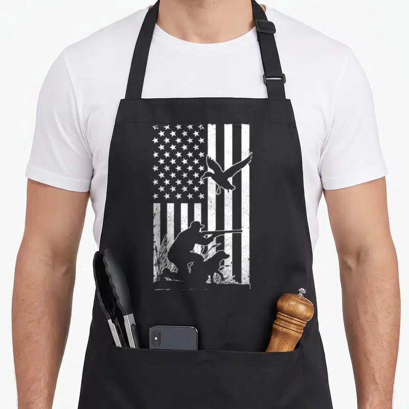 Flag cotton duck apron - Distressed USA flag background with white silhouette of kneeling hunter, dog, and flying duck — shown on a black cotton canvas apron against a white background
