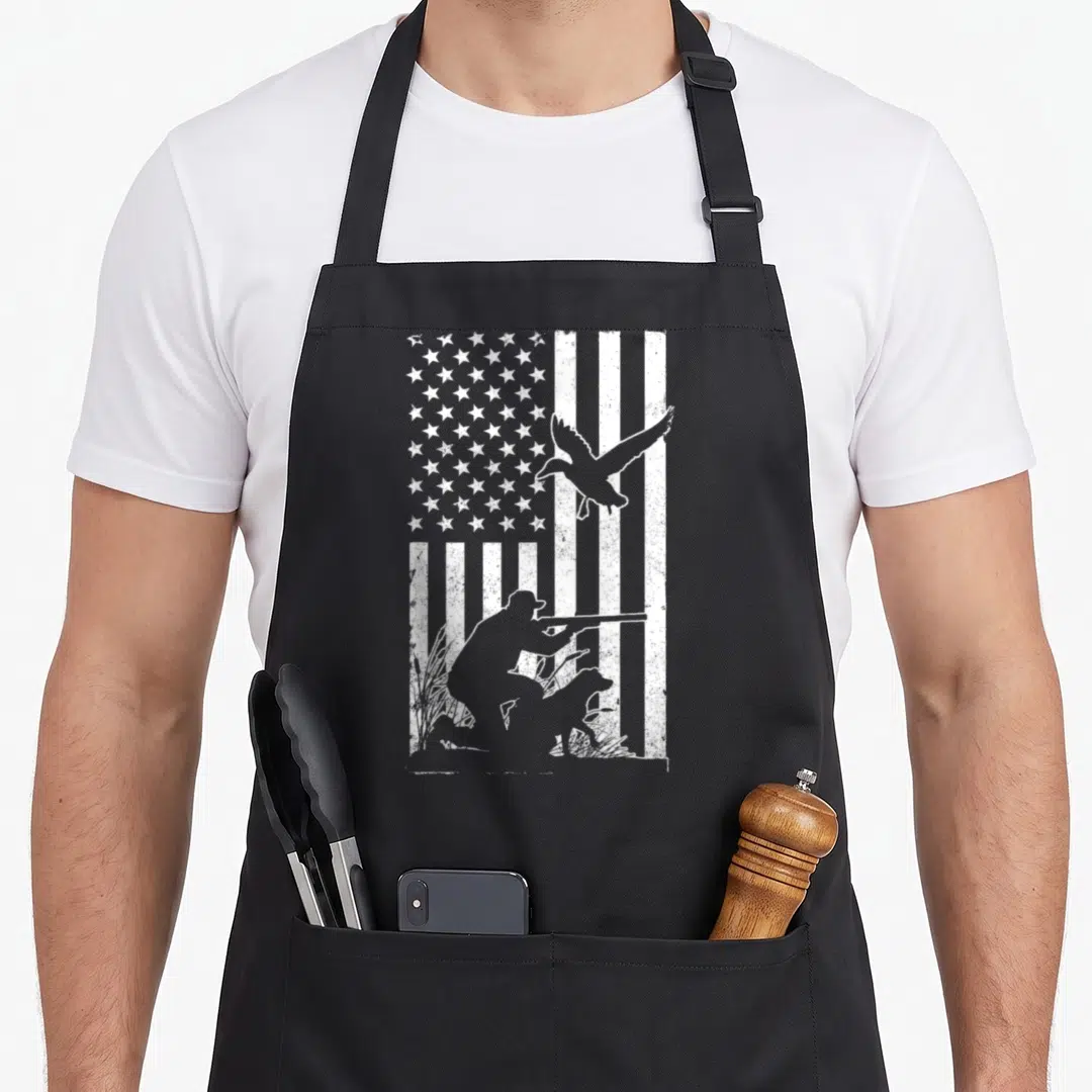 Flag cotton duck apron - Distressed USA flag background with white silhouette of kneeling hunter, dog, and flying duck — shown on a black cotton canvas apron against a white background