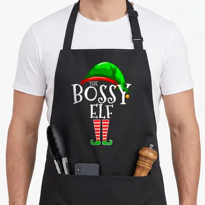 Bossy personalized family aprons - The Bossy Elf text with green hat and red striped legs cartoon — shown on a black cotton canvas apron against a white background
