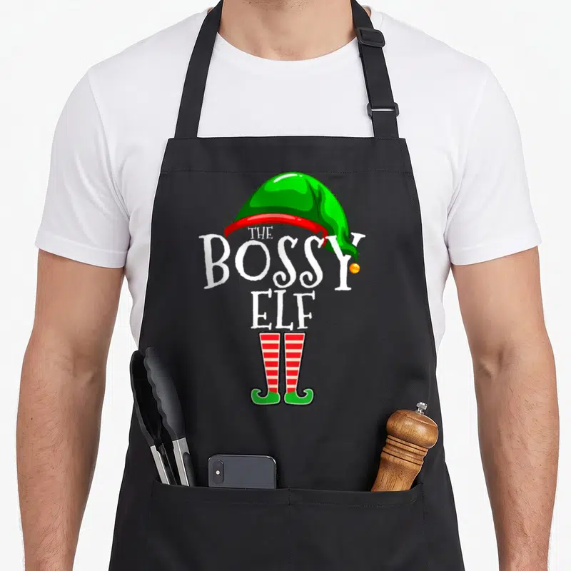 Bossy personalized family aprons - The Bossy Elf text with green hat and red striped legs cartoon — shown on a black cotton canvas apron against a white background