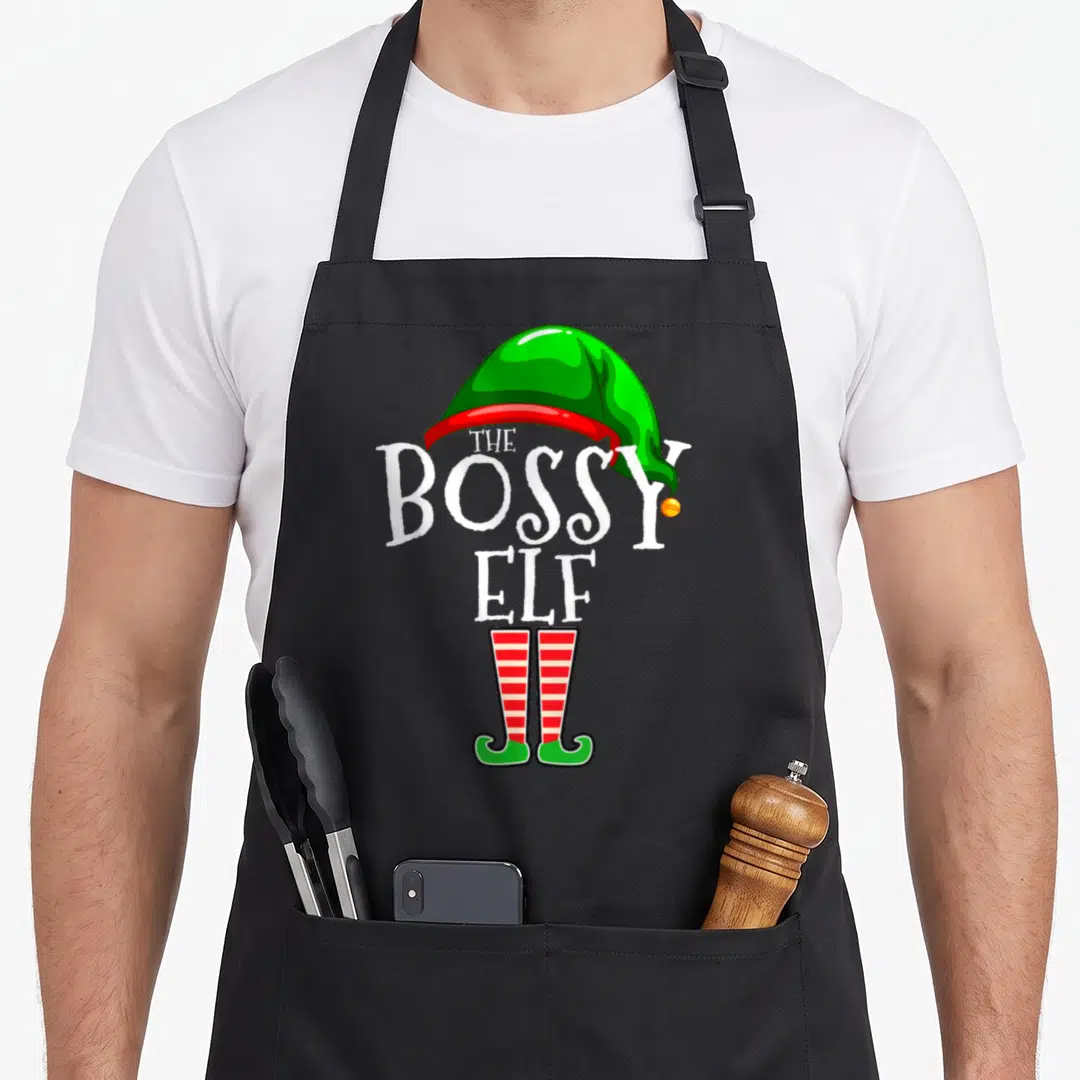 Bossy personalized family aprons - The Bossy Elf text with green hat and red striped legs cartoon — shown on a black cotton canvas apron against a white background