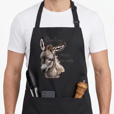 Smile donkey apron - Realistic donkey head illustration next to stacked brown text reading Donkeys Make me smile — shown on a black cotton canvas apron against a white background