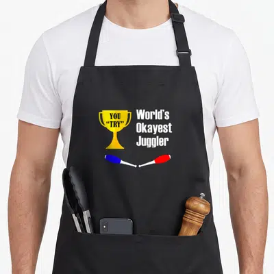 Okayest juggling apron - Gold trophy reading YOU TRY next to World's Okayest Juggler text above blue and red clubs — shown on a black cotton canvas apron against a white background