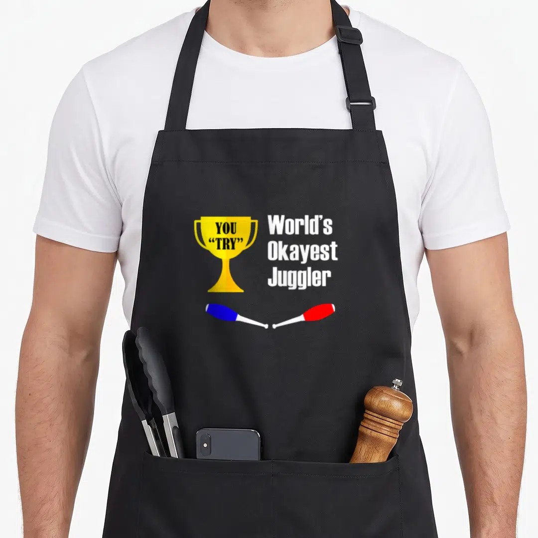 Okayest juggling apron - Gold trophy reading YOU TRY next to World's Okayest Juggler text above blue and red clubs — shown on a black cotton canvas apron against a white background