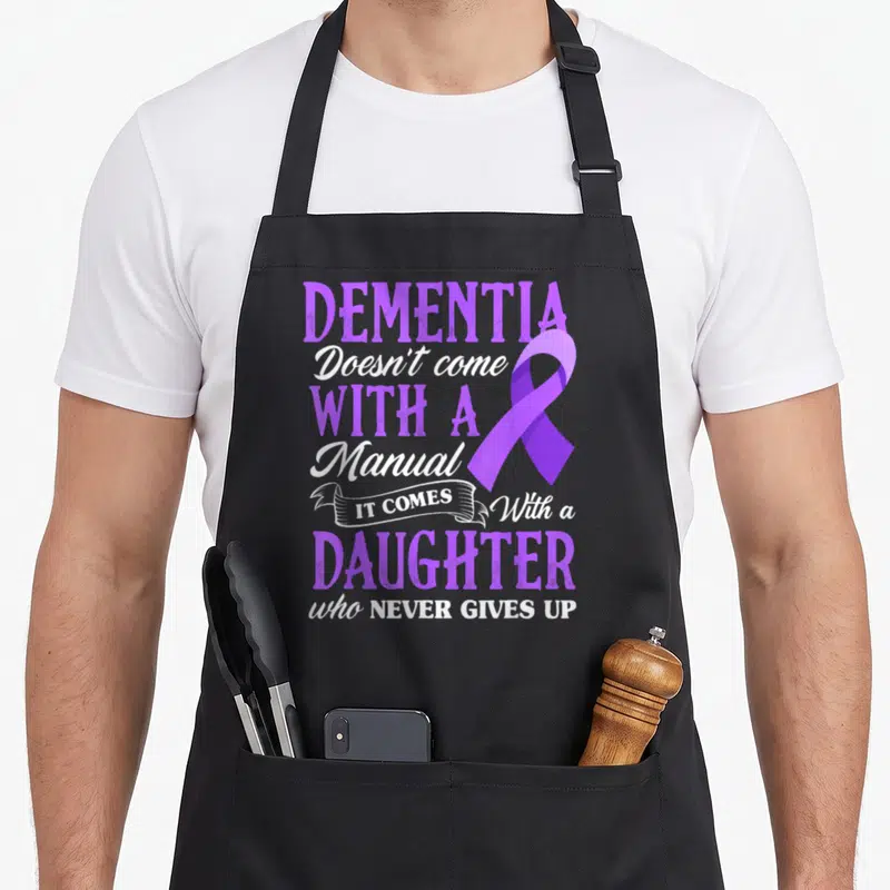 Dementia mom daughter aprons - Purple awareness ribbon overlapping vertical text reading DEMENTIA Doesn't come — shown on a black cotton canvas apron against a white background