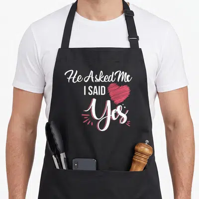 Asked proposal apron - Stacked He Asked Me I Said Yes text with pink sketch heart graphic — shown on a black cotton canvas apron against a white background