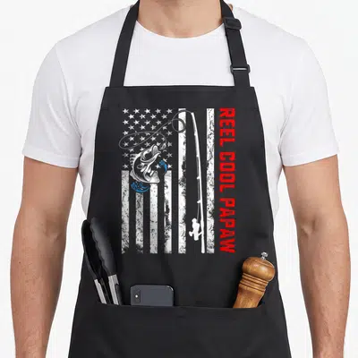 Reel papaw father day apron - Distressed US flag background with white fishing rod, jumping fish, and vertical red text — shown on a black cotton canvas apron against a white background