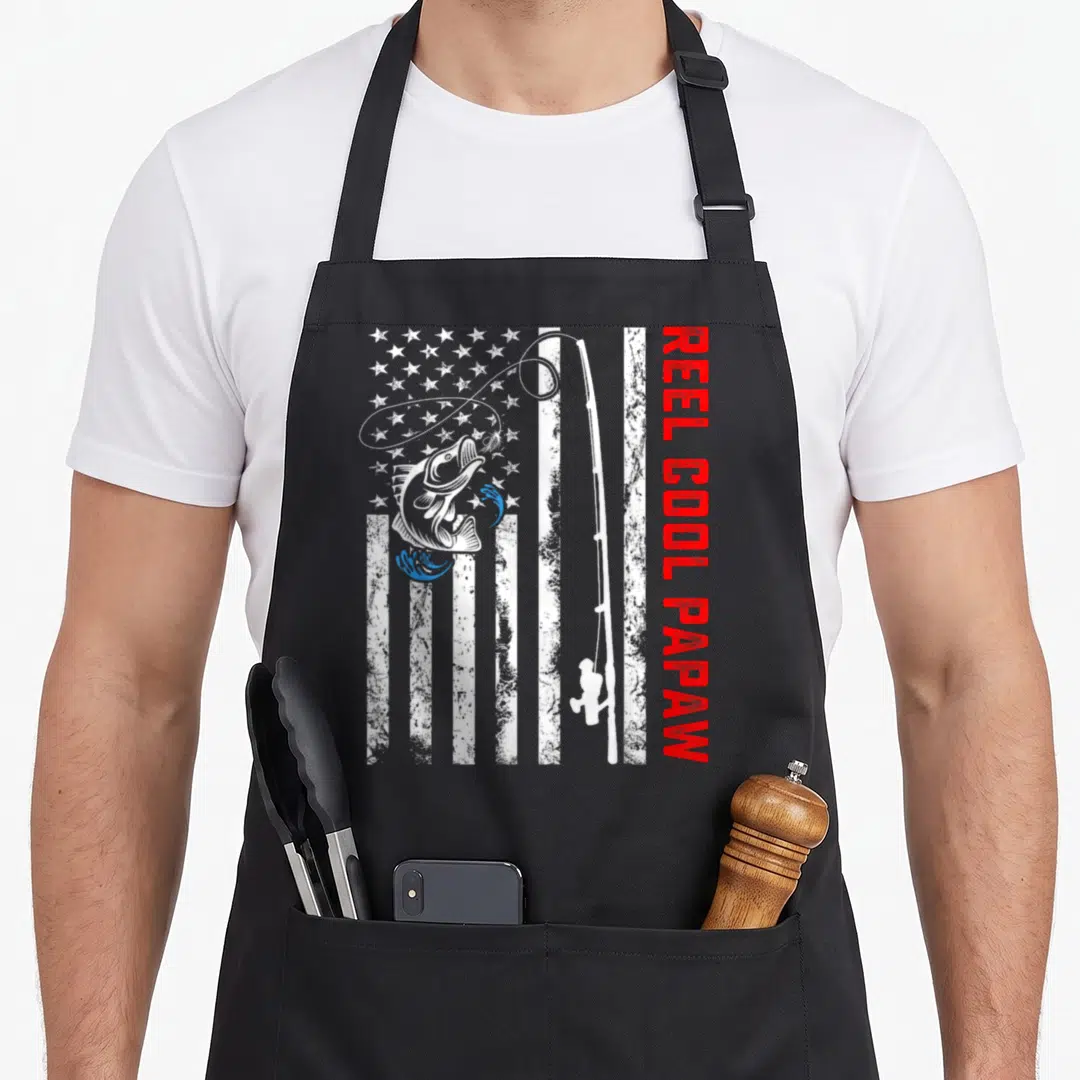 Reel papaw father day apron - Distressed US flag background with white fishing rod, jumping fish, and vertical red text — shown on a black cotton canvas apron against a white background