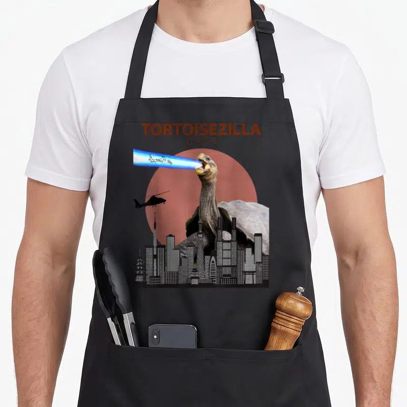 Tortoisezilla tortoise apron - Giant tortoise breathing blue beam over city skyline with brown text and red sun circle — shown on a black cotton canvas apron against a white background