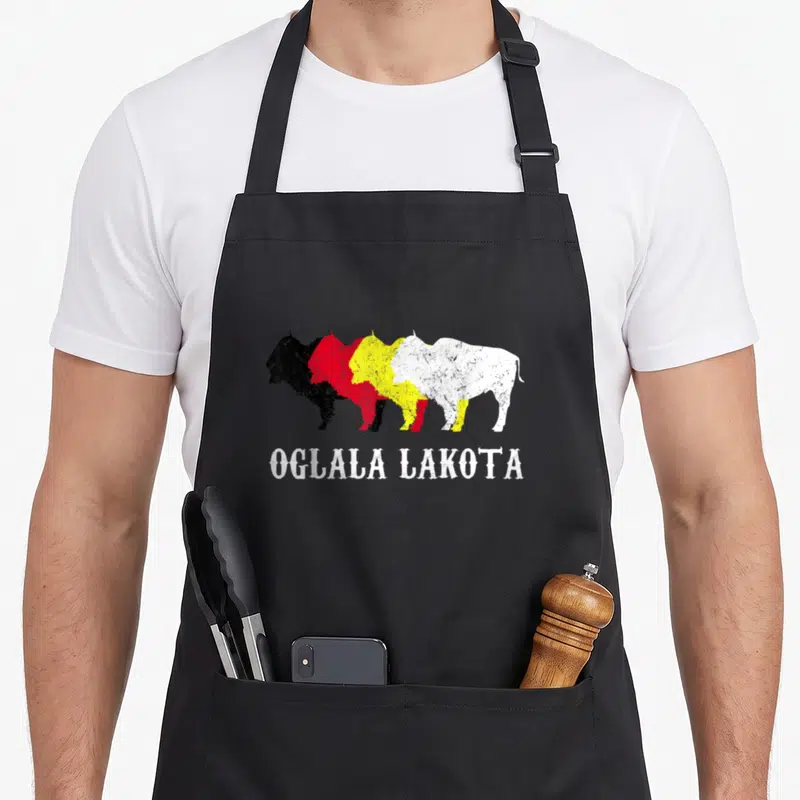 Oglala chiefs apron - Four overlapping bison in black red yellow white with OGLALA LAKOTA text below — shown on a black cotton canvas apron against a white background