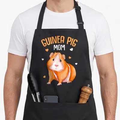 Pig mother's day aprons - Cartoon orange and white guinea pig with hearts and stacked text reading Guinea Pig Mom — shown on a black cotton canvas apron against a white background