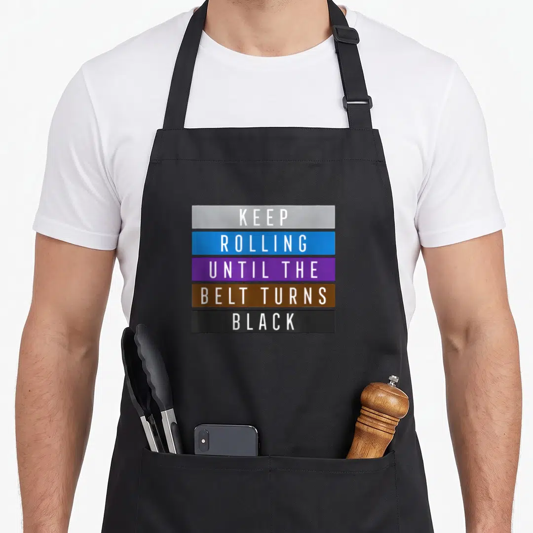 Rolling jitsu apron - Stacked white text on grey blue purple brown and black rectangles reading KEEP ROLLING UNTIL — shown on a black cotton canvas apron against a white background