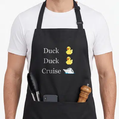 Cruise duck apron - Stacked text reading Duck Duck Cruise beside two yellow rubber ducks and a white cruise ship — shown on a black cotton canvas apron against a white background
