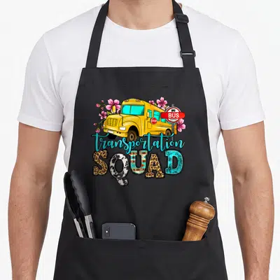 Transportation school bus apron - Yellow cartoon bus with pink flowers, bus stop sign, and patterned SQUAD text — shown on a black cotton canvas apron against a white background