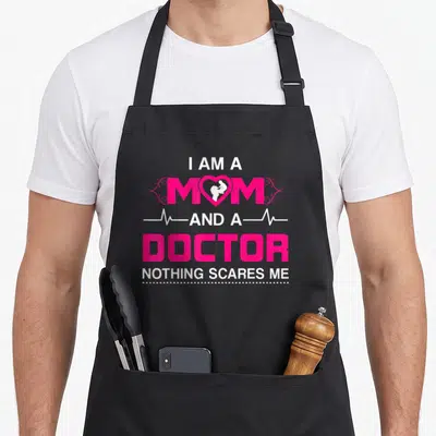 Scare doctor apron - Pink and white typography with mother child silhouette in heart and heartbeat line flanking text — shown on a black cotton canvas apron against a white background