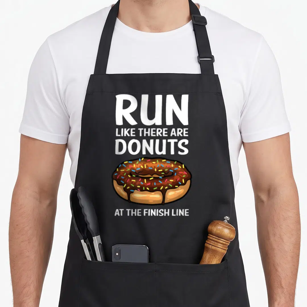 Running donut apron - Cartoon chocolate donut with white text reading Run Like There Are Donuts At The Finish Line — shown on a black cotton canvas apron against a white background