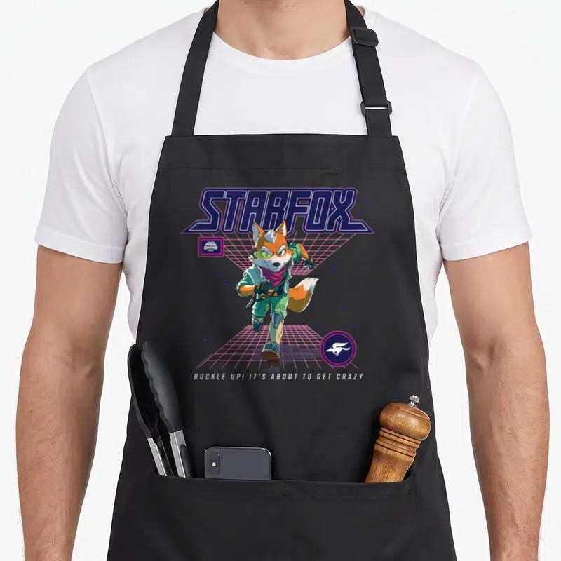 Galaxy novelty apron - Starfox lettering, orange fox on pink grid, buckle up text, retro style — shown on a black cotton canvas apron against a white background