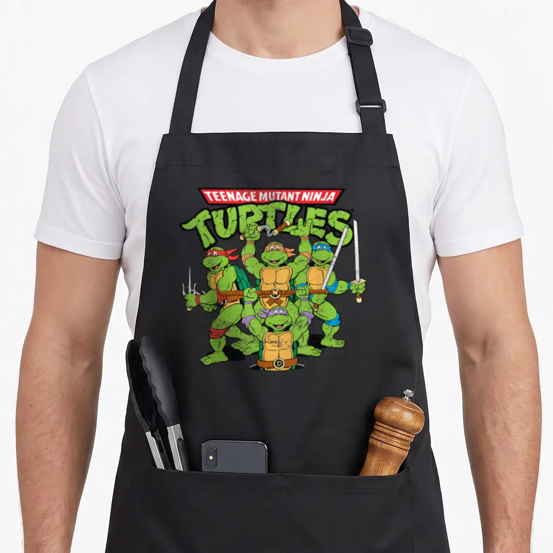 Vintage teenage cooking aprons - Four turtles with weapons under TEENAGE MUTANT NINJA TURTLES text in cartoon style — shown on a black cotton canvas apron against a white background