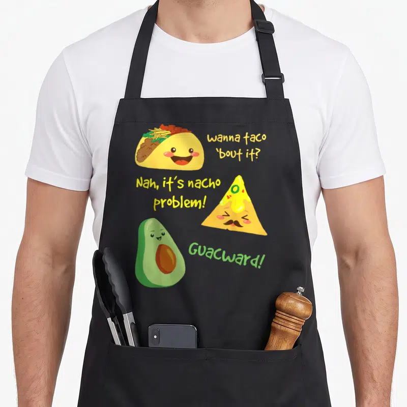 Taco food aprons - Cartoon taco, mustached nacho, and avocado with punny text in yellow and green — shown on a black cotton canvas apron against a white background