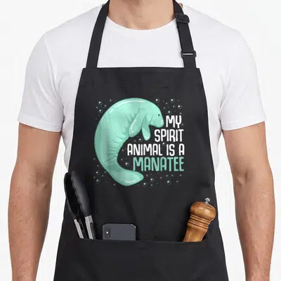 Sea cow apron - Cartoon mint green manatee curving around white text My Spirit Animal is a Manatee with bubbles — shown on a black cotton canvas apron against a white background