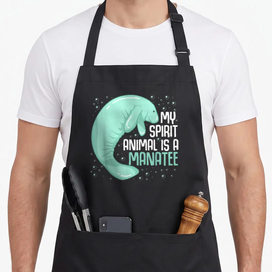 Sea cow apron - Cartoon mint green manatee curving around white text My Spirit Animal is a Manatee with bubbles — shown on a black cotton canvas apron against a white background