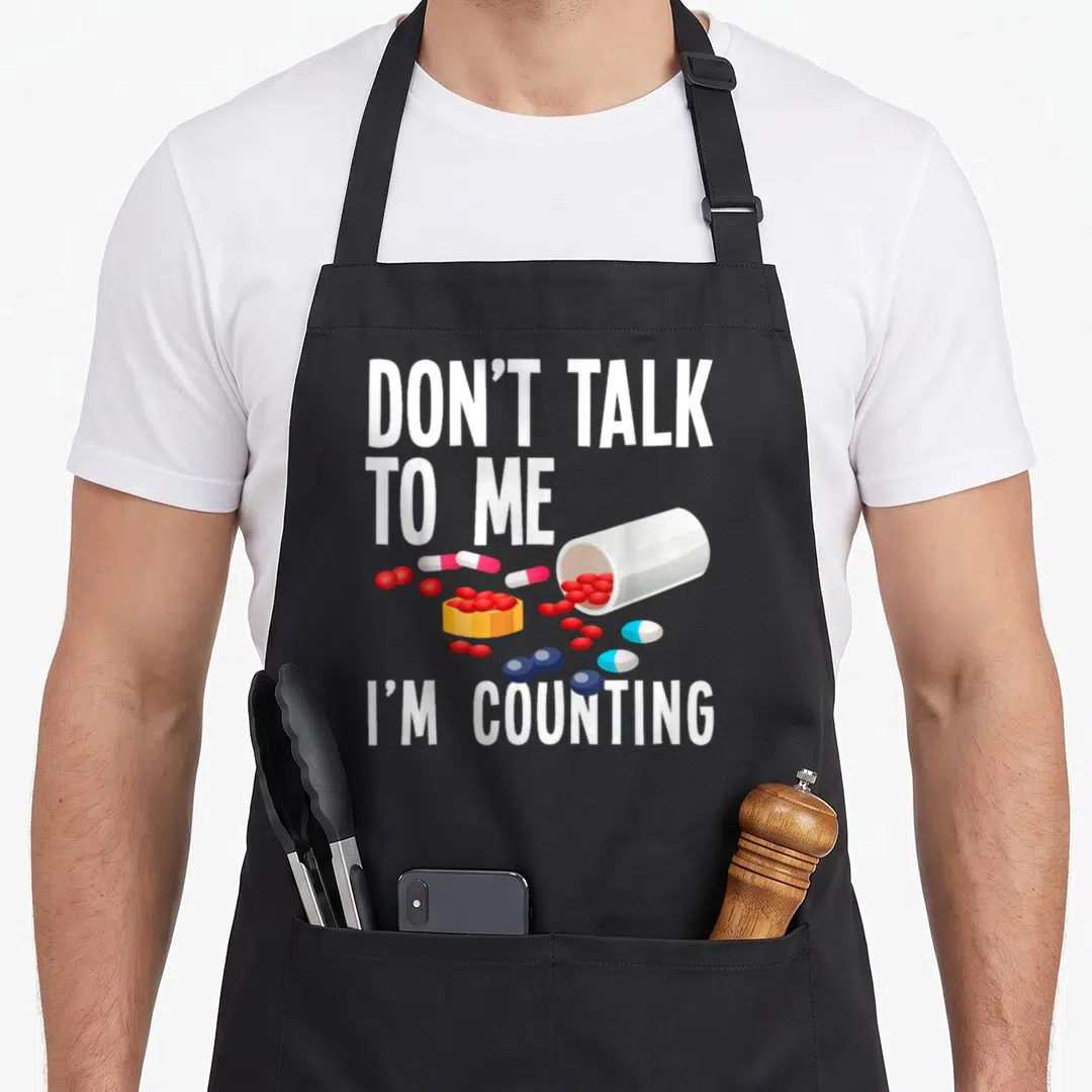 Counting pharmacy apron - DON'T TALK TO ME I'M COUNTING text above spilled red blue and yellow pills — shown on a black cotton canvas apron against a white background