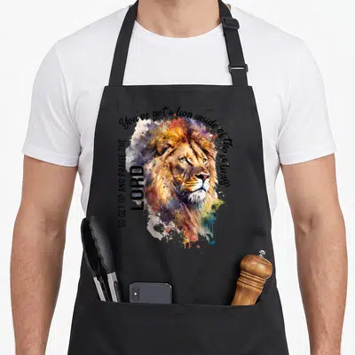 Inside christian apron - Watercolor lion head with orange blue purple splashes and praise text on black background — shown on a black cotton canvas apron against a white background