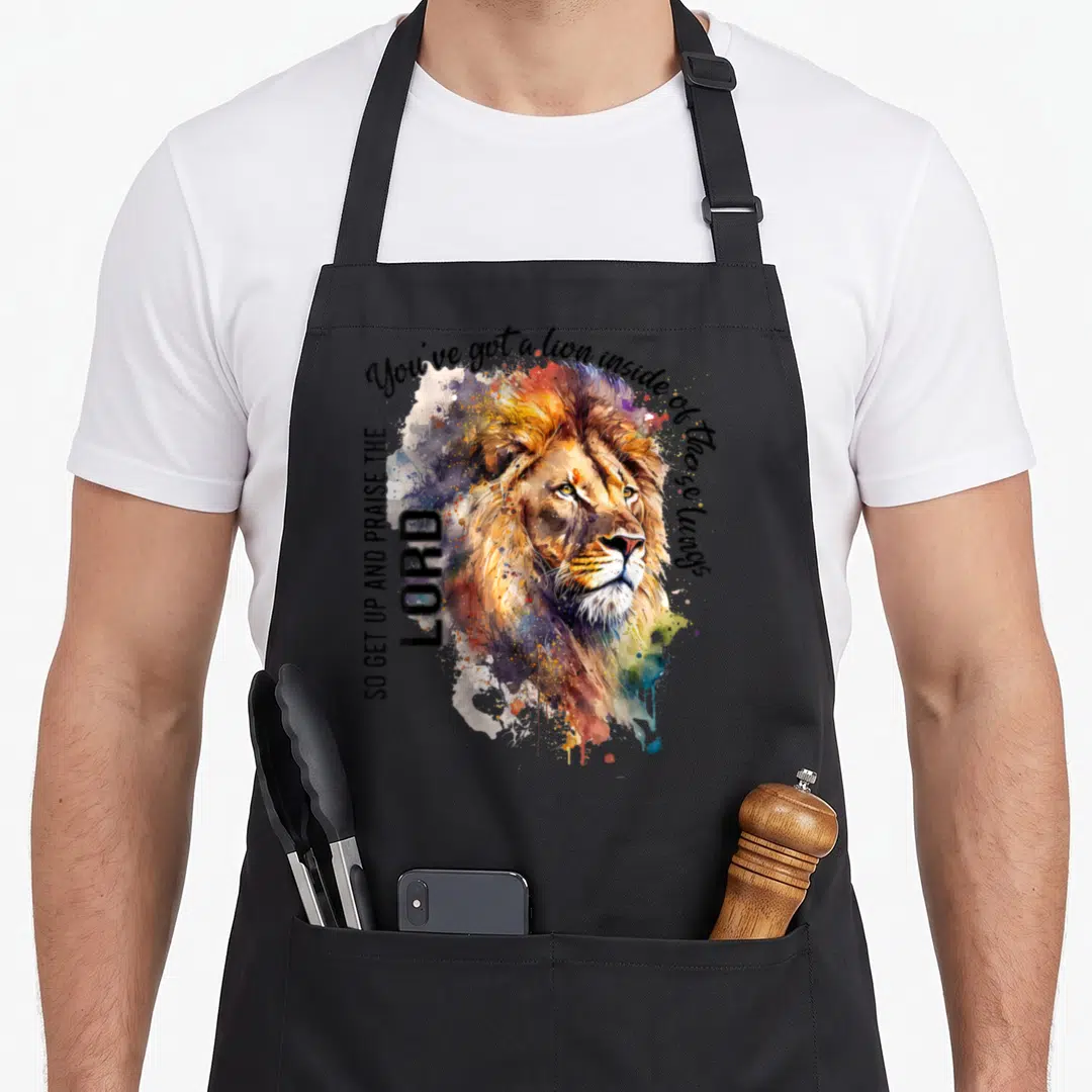 Inside christian apron - Watercolor lion head with orange blue purple splashes and praise text on black background — shown on a black cotton canvas apron against a white background