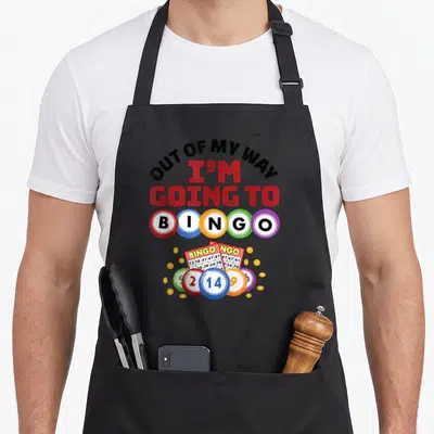 Way bingo apron - OUT OF MY WAY I'M GOING TO BINGO text with colorful balls and cards — shown on a black cotton canvas apron against a white background