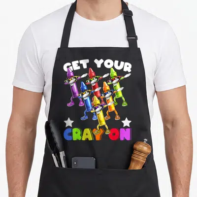 Cray crayons apron - Six dabbing crayons in rainbow colors under GET YOUR CRAYON text with white stars — shown on a black cotton canvas apron against a white background