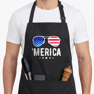 Fourth american flag apron - Aviator sunglasses with US flag lenses above distressed 'MERICA text and five stars — shown on a black cotton canvas apron against a white background