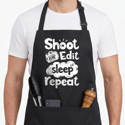 Sleep photographer apron - Shoot Edit Sleep Repeat text with camera icon and sleeping cloud in distressed white font — shown on a black cotton canvas apron against a white background