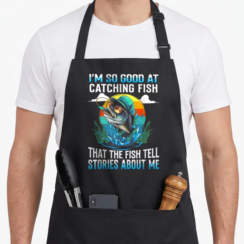 Catching fish apron - Cartoon fish in sunglasses with striped sun and fishing quote — shown on a black cotton canvas apron against a white background