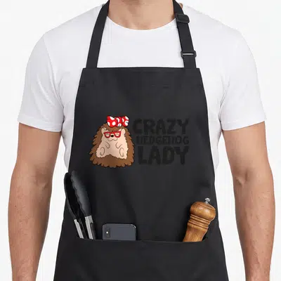Comfort crazy hedgehog lady apron - Cartoon brown hedgehog with red glasses and polka dot bow next to stacked black text — shown on a black cotton canvas apron against a white background